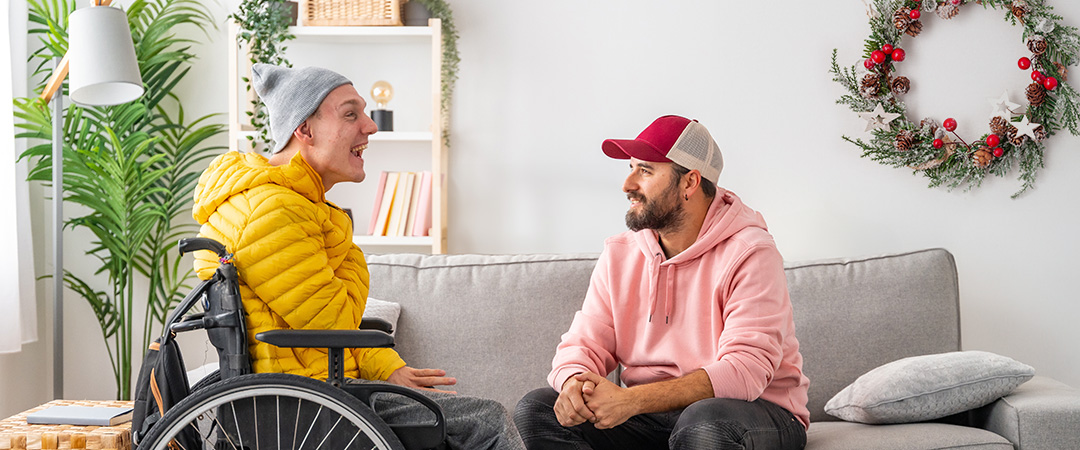 A man in a wheelchair talking with a man sitting on a sofa.
